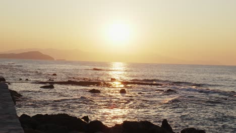 Toma-En-Cámara-Lenta-De-La-Puesta-De-Sol-Sobre-Las-Olas-Del-Océano-Chocando-Contra-Las-Piedras,-Hermoso-Fondo-Del-Mar-Egeo-En-Chania,-Creta-Grecia