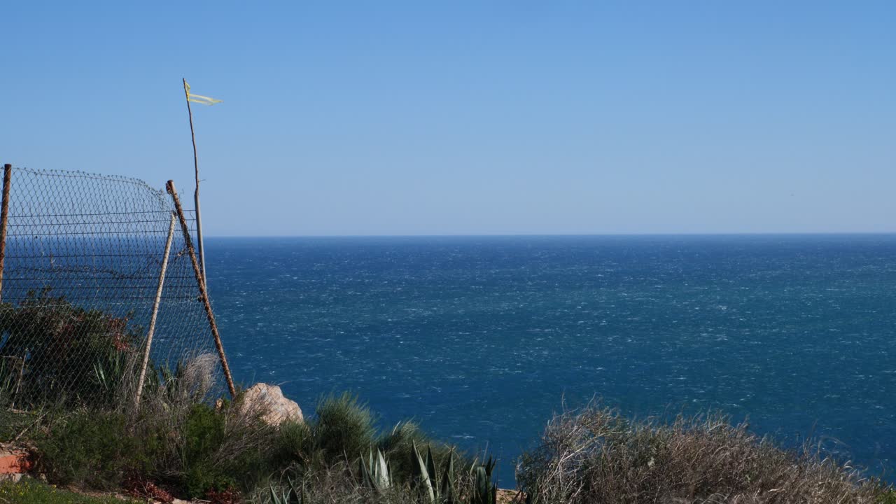 Arbusti che ondeggiano con il vento forte, accanto a una recinzione. Il Mar Mediterraneo sullo sfondo.