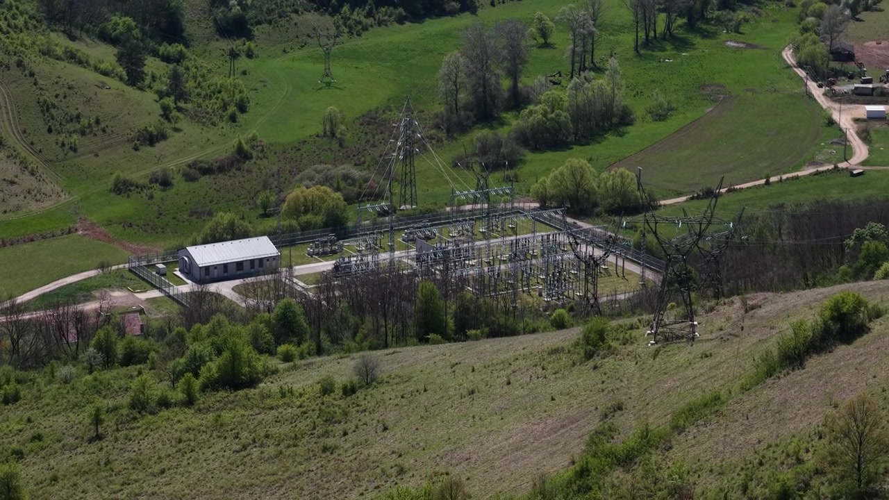 Pohled shora na elektrickou rozvodnu a přenosové stožáry distribuující energii napříč svěží zelenou venkovskou krajinou.