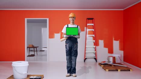 cuerpo lleno de pintor hombre asiático con casco de seguridad sonriendo y mostrando portátil de pantalla verde a la cámara mientras está de pie con la pared de pintura en la habitación