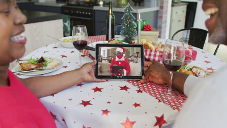 pareja afroamericana con vino usando tableta para la videollamada de navidad con santa en la pantalla
