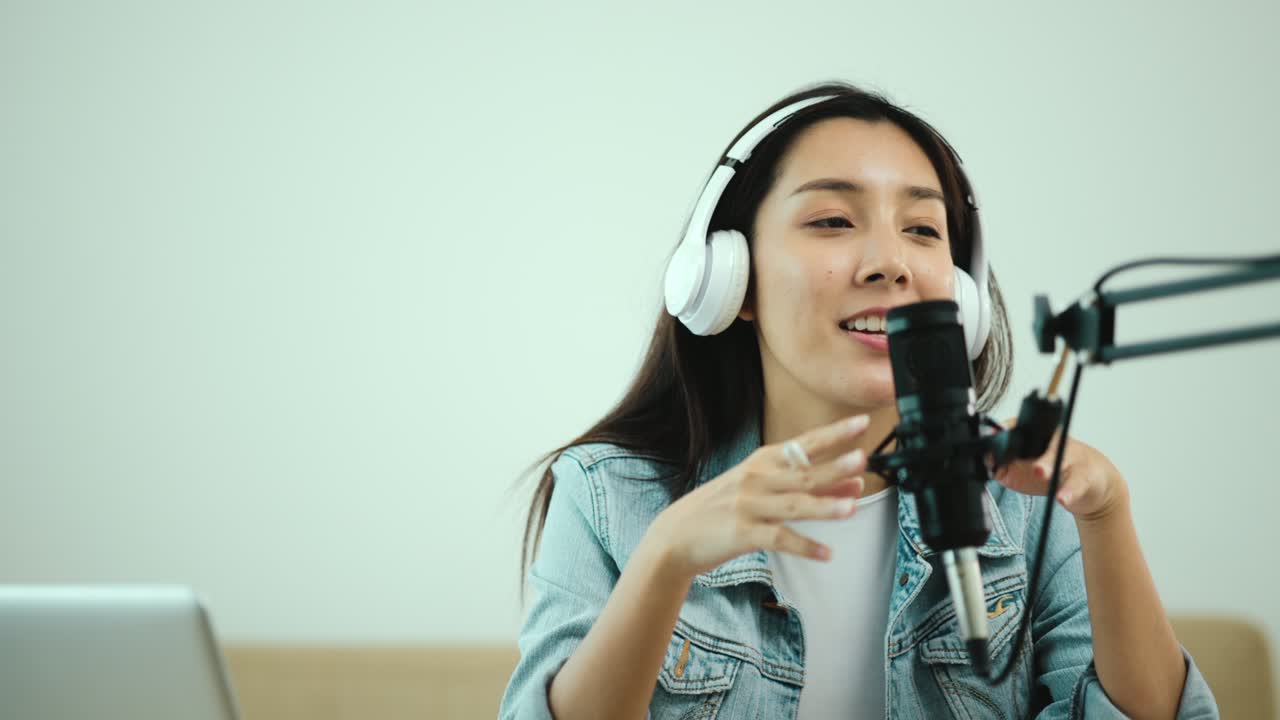 컨텐츠 제작자 여성 호스트는 헤드폰과 마이크로 노트북에서 캐스트를 스트리밍하고 방송 스튜디오에서 게스트 대화를 응원합니다.