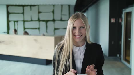 cheerful young woman with phone in office. pretty blond woman with long hair wearing elegant black suit and smiling charmingly at camera inside of modern office