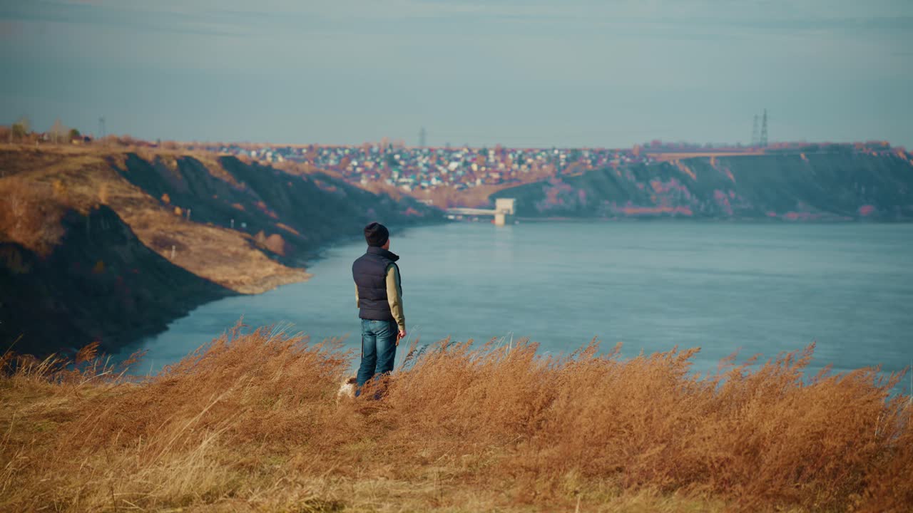 En ensom skikkelse betragter det fredfyldte landskab fra en græsklædt klippe og skuer ud over det rolige vand nedenfor, omgivet af blidt efterårsløv og fjerne boligområder i et fantastisk panorama
