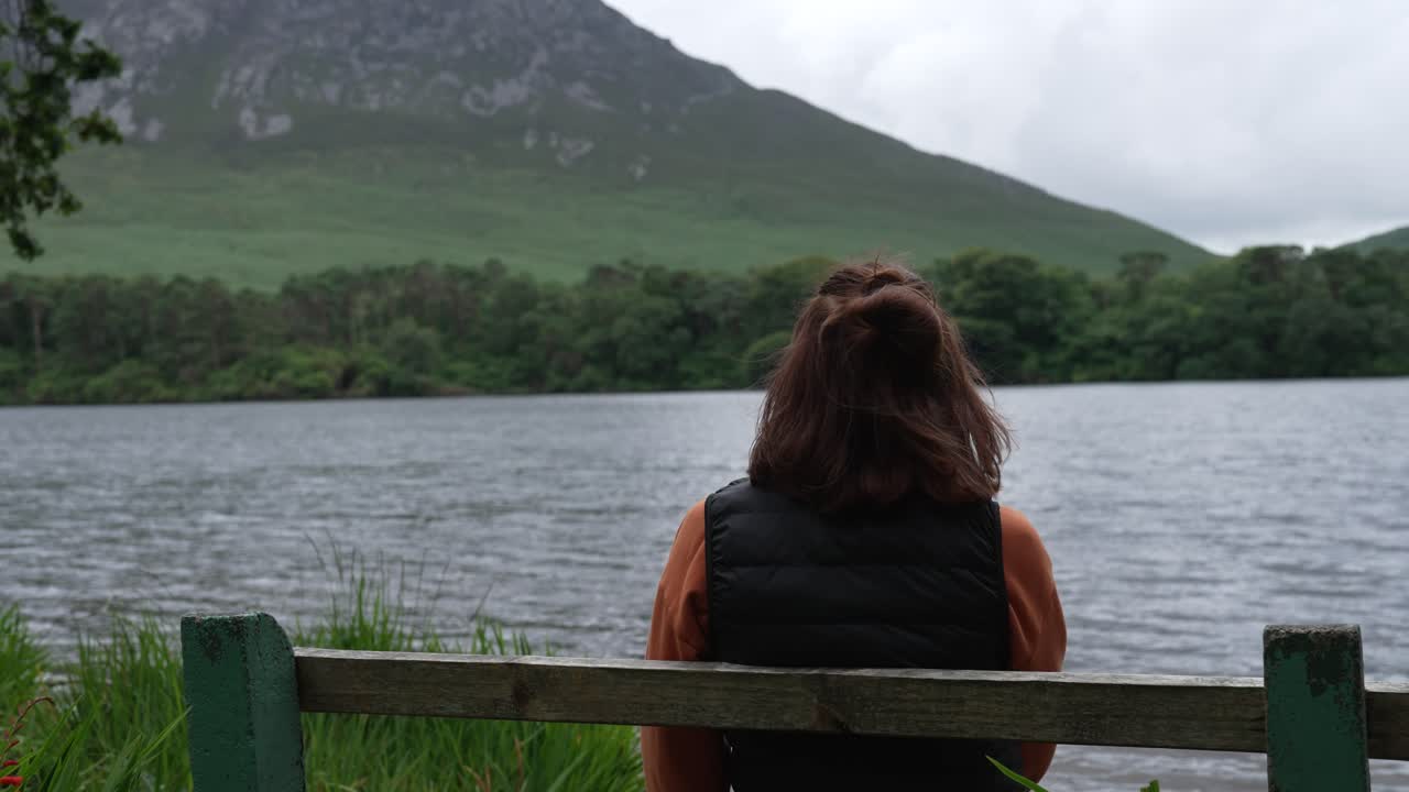 Foto da dietro: una donna seduta tranquillamente su una panchina, mentre ammira un meraviglioso lough irlandese e una catena montuosa. Un momento di quieta contemplazione e connessione con la natura.