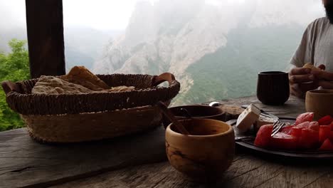 Köstliches-Traditionelles-Frühstück-Auf-Handgefertigtem-Holztisch-Auf-Der-Cottage-Terrasse-Mit-Geschnittenem-Frischem-Brot,-Käse,-Tomate,-Tee,-Kaffee-Heißgetränk-Und-Grüner-Bergfelsen-Waldnebellandschaft