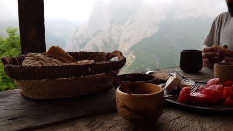 delicioso desayuno tradicional en una mesa de madera hecha a mano en la terraza de la cabaña con pan fresco en rodajas, queso, tomate, té, café, bebida caliente y paisaje de niebla del bosque de pradera de roca de montaña verde