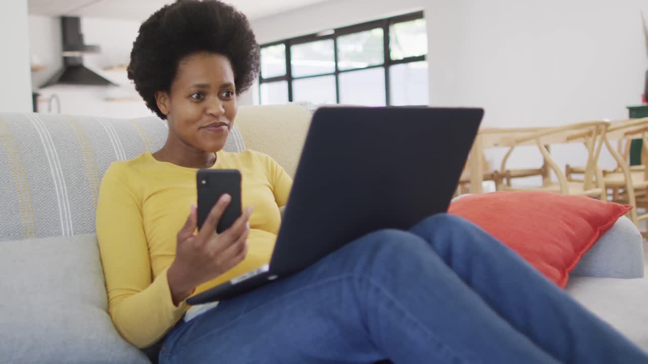 Happy african american woman sitting on sofa using laptop and smartphone