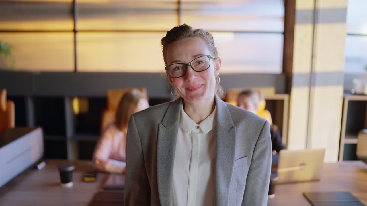 Portrait of a happy and confident middle-aged blonde girl in glasses in a gray business suit who is posing near the table in a modern sunny office