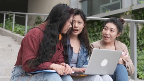 group of asian female students using a laptop and studying together while sitting on the stairs of the university campus.