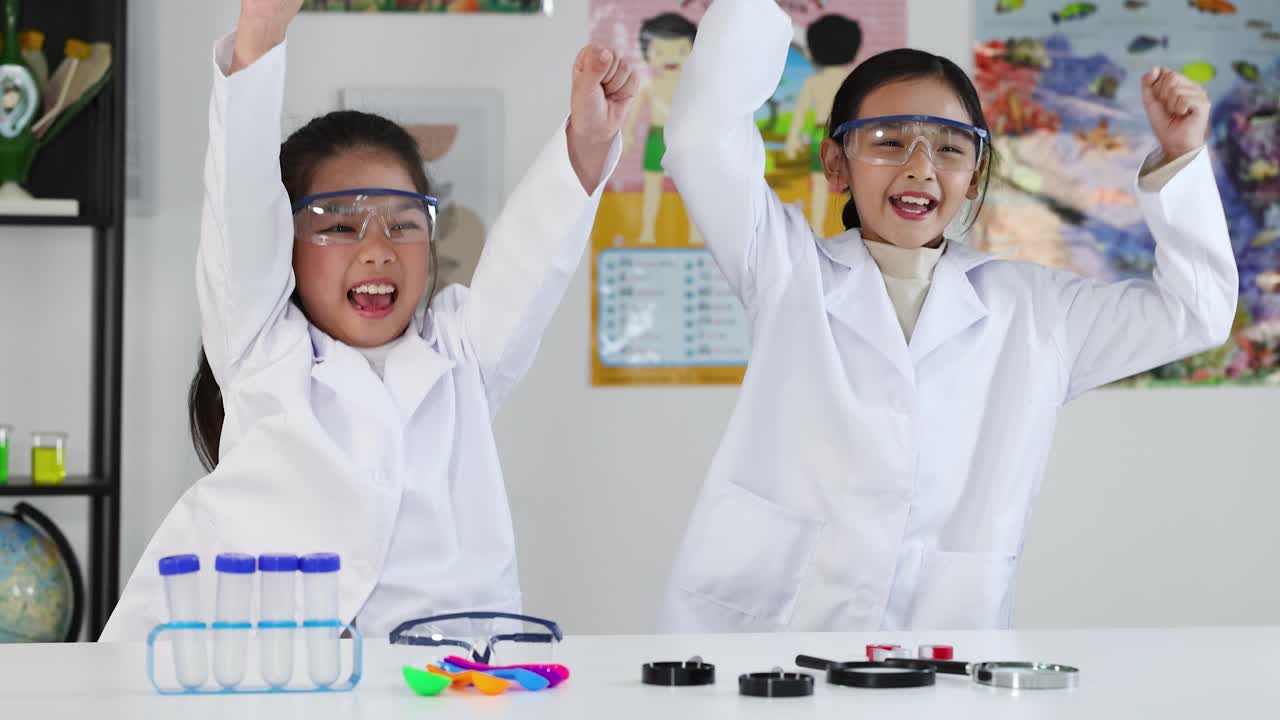 Dua gadis muda Asia mengenakan jas lab dan kacamata pelindung bersorak gembira di ruang kelas sains.