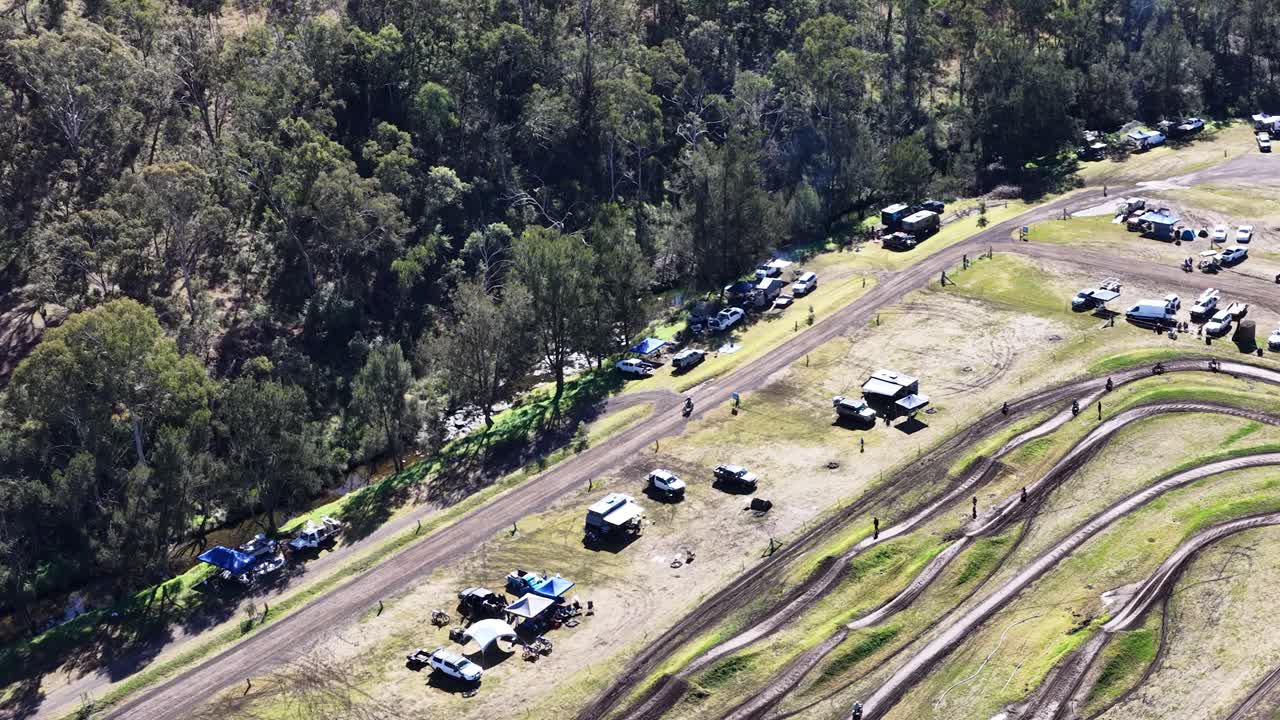 Drone footage captures motocross track, parked vehicles, and forest edge in bright daylight conditions