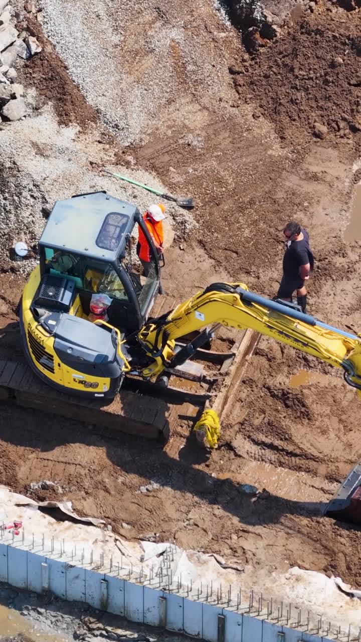 Droneoptagelse fra høj vinkel af en gul gravemaskine, der graver mudder nær en betonstøttemur.