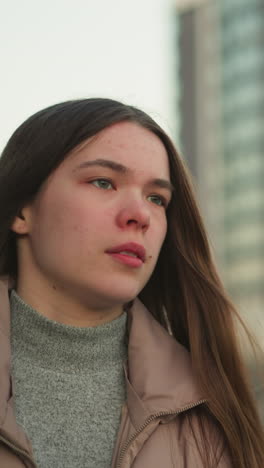 un primer plano frontal de una joven con una chaqueta de melocotón y un suéter interior gris. su boca está ligeramente abierta, y su expresión parece pensativa, con el telón de fondo de edificios altos