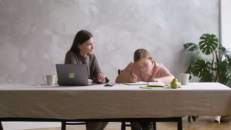 boy with down syndrome doing homeworks sitting at table in the living room at home. his mother helps him