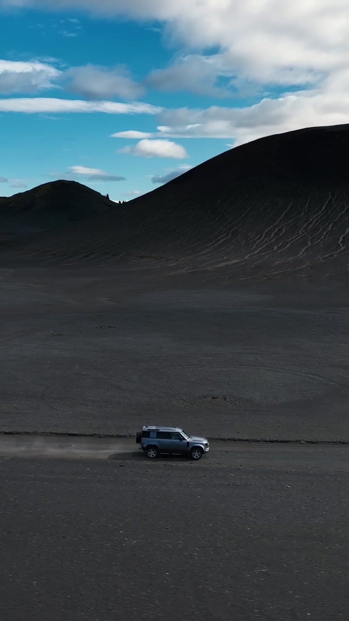 Drone footage of car traveling across Iceland highlands, volcanic desert, and striking rugged mountains. Vertical side view.