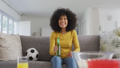 Afroamerikanerin-Sitzt-Zu-Hause-Auf-Einem-Sofa-Im-Wohnzimmer,-Schaut-Sich-Sport-Im-Fernsehen-An-Und-Trinkt