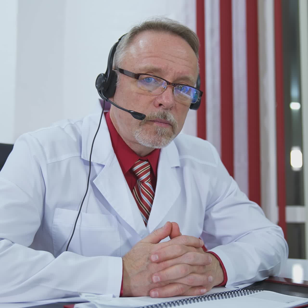 Serious doctor giving audio consultation call. Professional senior therapist sitting in medical office with headset and talking. Remote consultation.