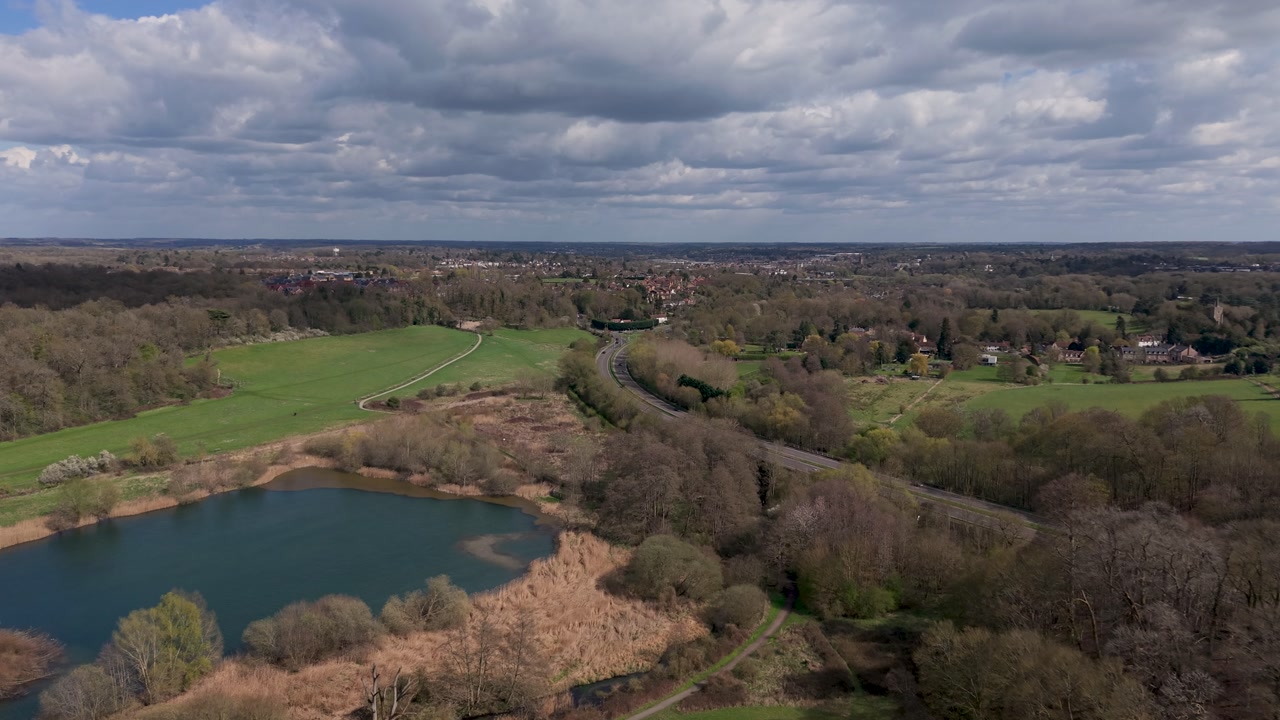 Drone flight over Panshanger Park, Osprey Lake on the left and Hertford ahead. Bright cloudy spring day shows green fields, woodland and walking paths in Hertfordshire, UK.