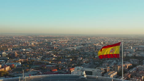 Volar-Alrededor-De-La-Bandera-Española-Ondeando-En-El-Viento-En-La-Azotea-Del-Rascacielos-De-Negocios.-Reveladora-Vista-Aérea-Del-Paisaje-Urbano-Iluminado-Por-El-Sol-Poniente.