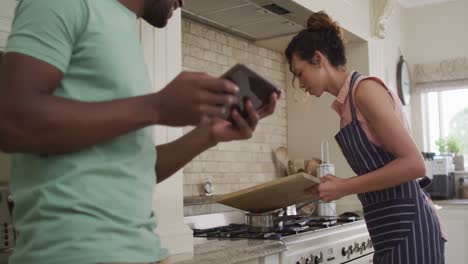 Happy-biracial-woman-putting-vegetables-into-saucepan-and-talking-to-her-partner-holding-tablet