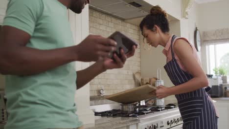 Feliz-Mujer-Birracial-Poniendo-Verduras-En-Una-Cacerola-Y-Hablando-Con-Su-Pareja-Sosteniendo-Una-Tableta