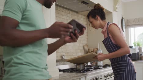 feliz mujer biracial poniendo verduras en la sartén y hablando con su pareja sosteniendo la tableta