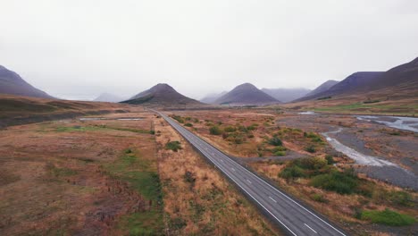 Aérea:-Revelación-Inversa-De-Un-SUV-Blanco-Que-Viaja-A-Lo-Largo-De-La-Carretera-De-Circunvalación-De-Islandia,-Que-Es-Una-Carretera-Panorámica-A-Través-De-Una-Pintoresca-Y-Remota-Zona-De-Fiordos-Que-Conduce-A-Picos-Gemelos-Con-Niebla-Y-Neblina-En-La-Distancia.