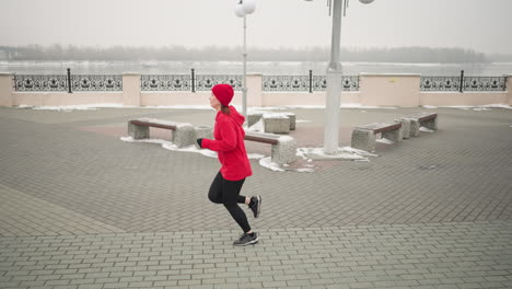 vista lateral de una mujer con capucha roja corriendo al aire libre durante el invierno en el pavimento entrelazado, paisaje urbano con bancos, barandillas decorativas, suelo cubierto de nieve y atmósfera serena cerca del río