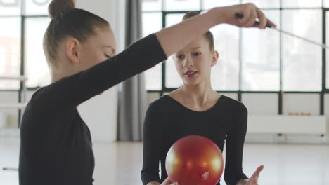 dos chicas rubias gimnásticas jugando con una banda y una pelota mientras hablan con su amiga antes de comenzar la clase de ballet