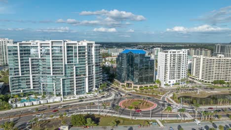 aerial hyperlapse of downtown sarasota, featuring a panning and orbiting view of the cityscape, roundabout, and heavy traffic under sunny skies with puffy clouds