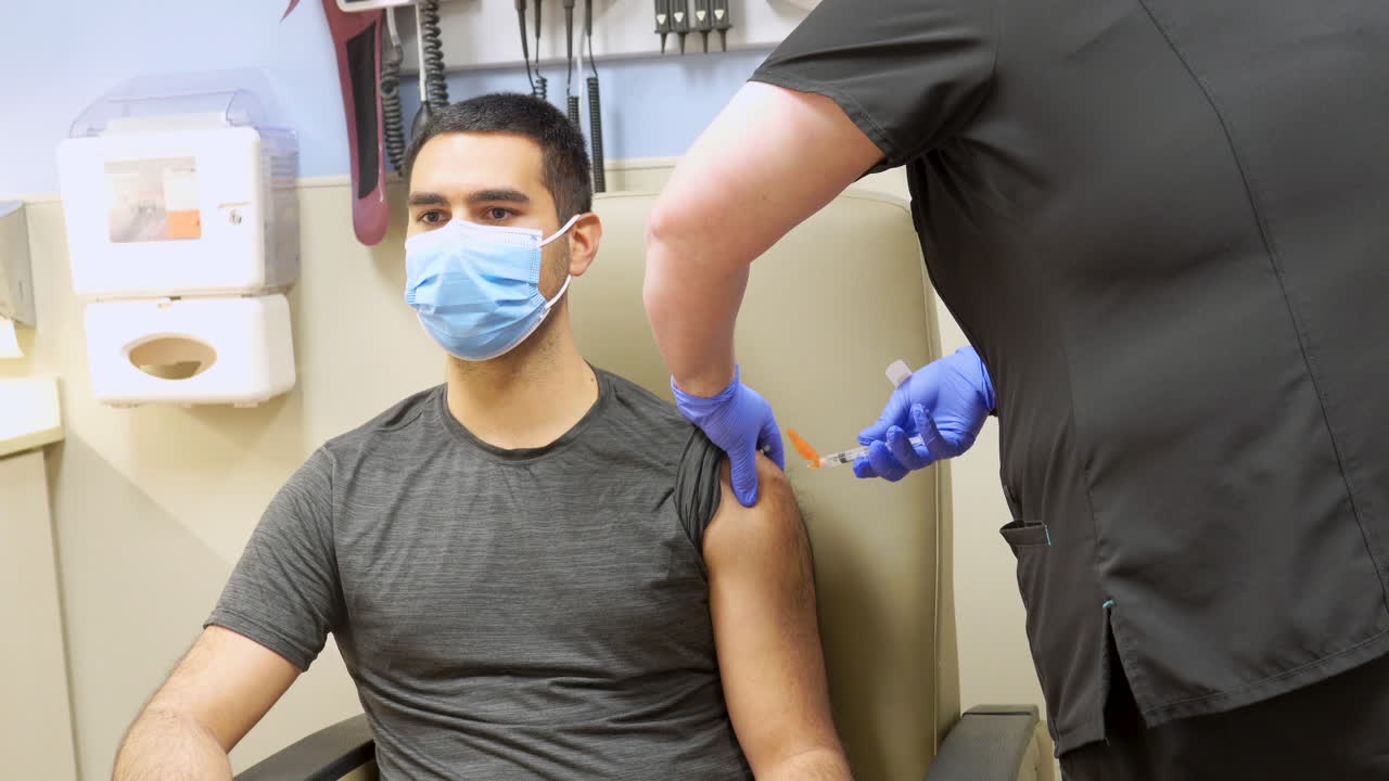 vacuna covid 19 inyectada en el brazo de un joven adulto de piel oscura de etnia minoritaria con una máscara médica, inyección de virus corona administrada por una enfermera en el consultorio del médico con el paciente sentado
