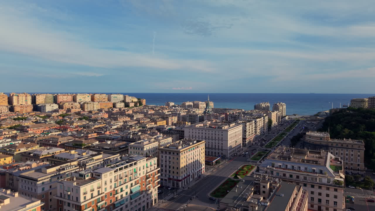 Drone forward shot crossing a central avenue with medians and greenery, showing residential blocks, the coastline, and blue sky in Genoa