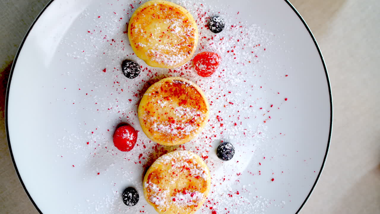 Delicious syrniki, traditional Eastern European cheese pancakes, garnished with powdered sugar and berries, rotate slowly on a white plate against a minimalist backdrop
