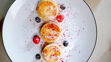 delicious syrniki, traditional eastern european cheese pancakes, garnished with powdered sugar and berries, rotate slowly on a white plate against a minimalist backdrop