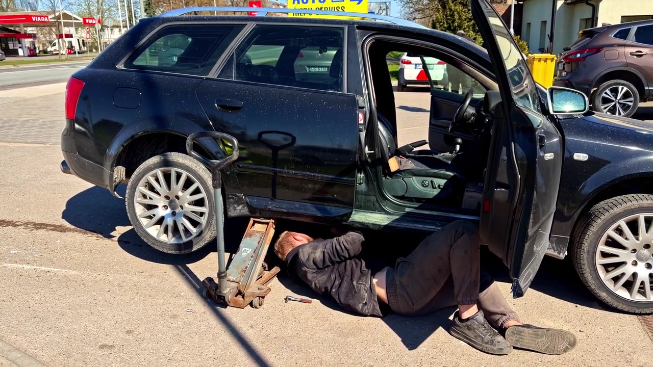 Mecânico trabalhando debaixo de um carro levantado em uma área de reparação automotiva ao ar livre em um dia ensolarado.