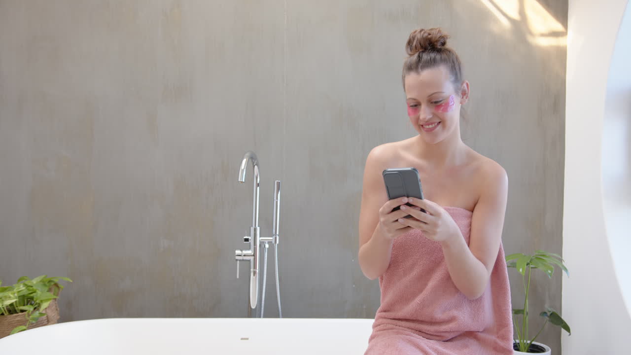 Using smartphone, woman in towel smiling and relaxing in bathroom, copy space
