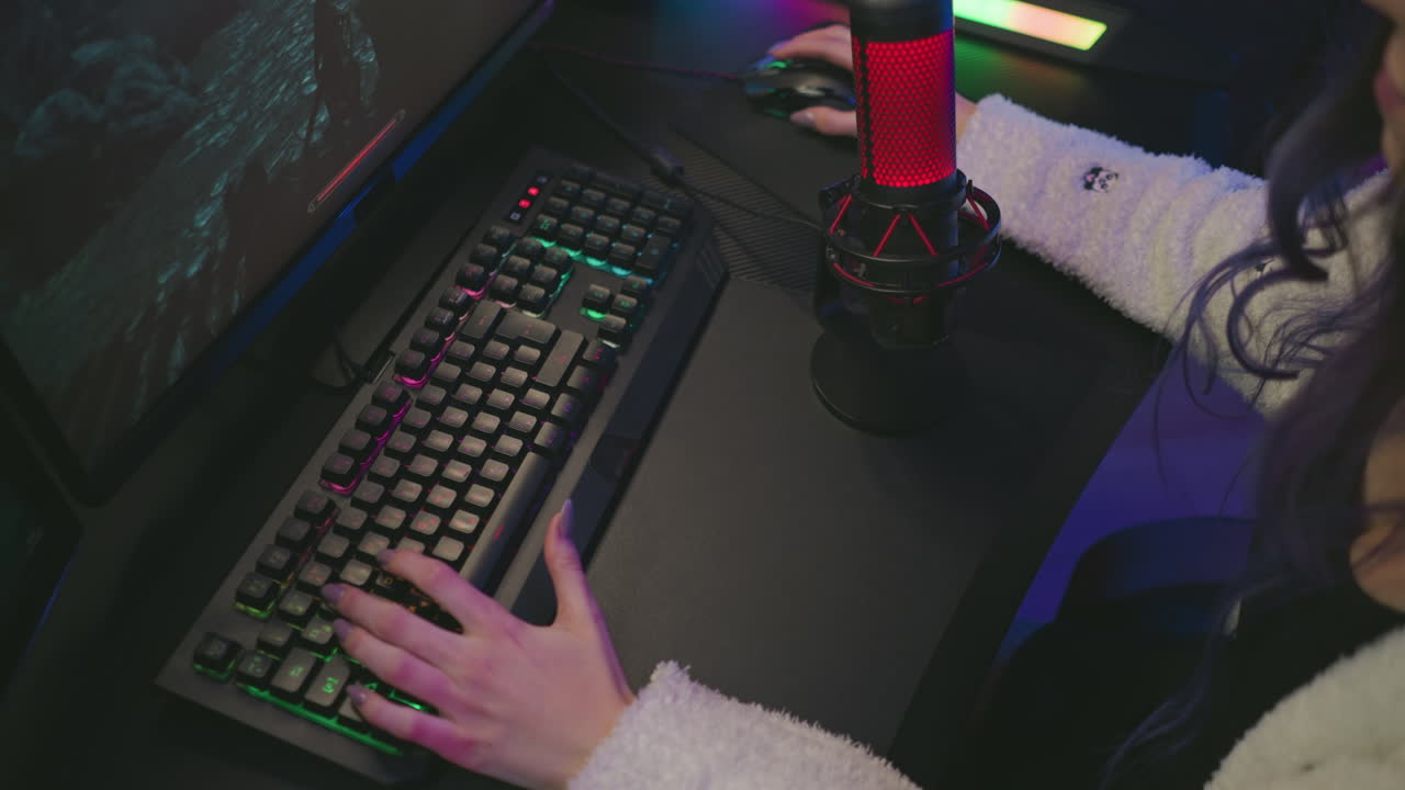 aerial view of woman in cozy sweater using colorful mechanical keyboard and gaming mouse to play video game on monitor with red microphone on desk inside illuminated modern studio setup
