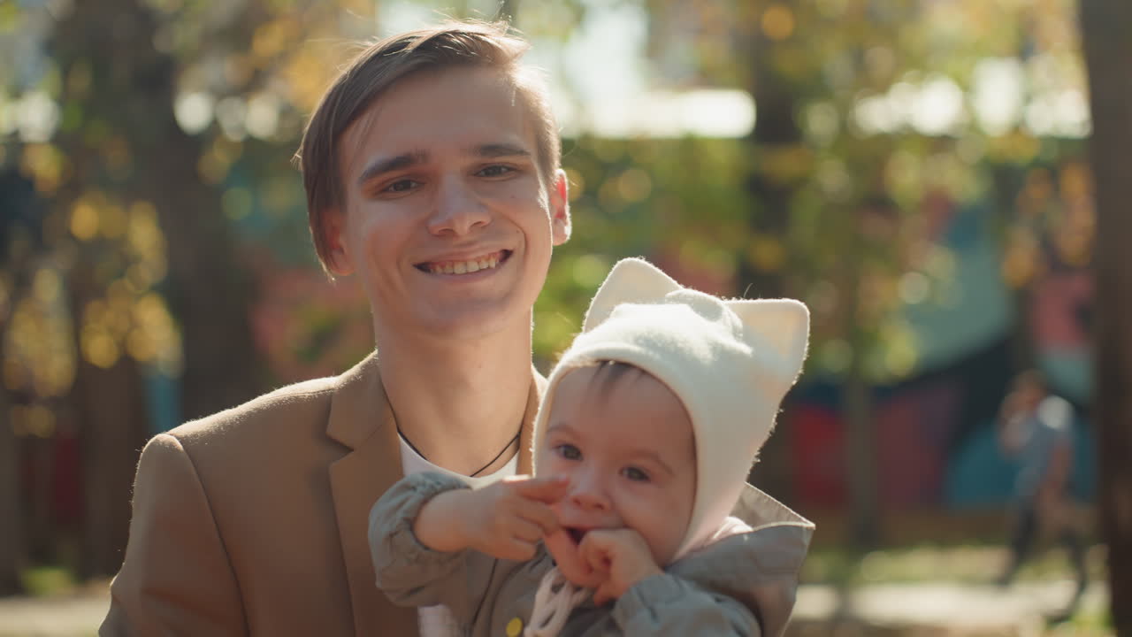 Ung, hvid far smiler med baby i parken. Baby tygger på bidering, hyggelig hue og varm frakke, gyldent sollys og vægmaleri som baggrund, kærlig forbindelse og familieglæde med ægteskabstema.