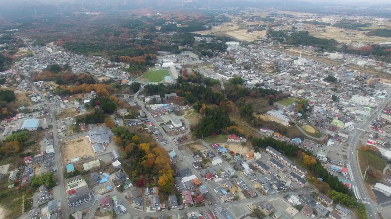 日本の福島住宅街を空撮。紅葉した木々、家々、そして遠くに見える風力タービン。