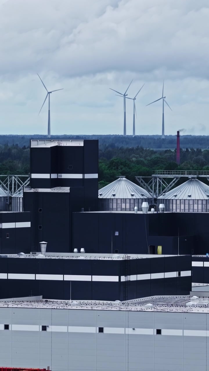 Industrial plant with wind turbines in distance highlighting energy and manufacturing, aerial ascend reveal vertical