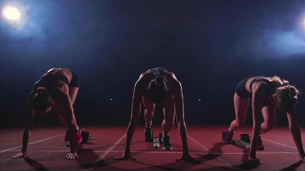 Female runners at athletics track crouching at the starting blocks before a race. In slow motion.