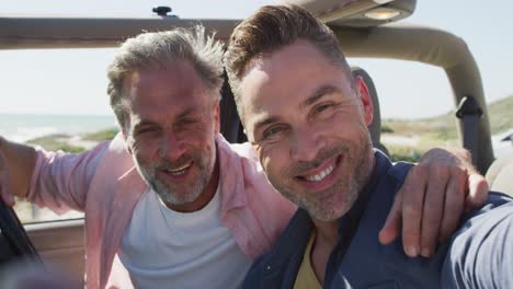 happy caucasian gay male couple taking selfies sitting in car smiling on sunny day at the beach