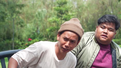 two cheerful and friendly asian university students are sitting on the bench in a campus park, waving their hands to calling their friends