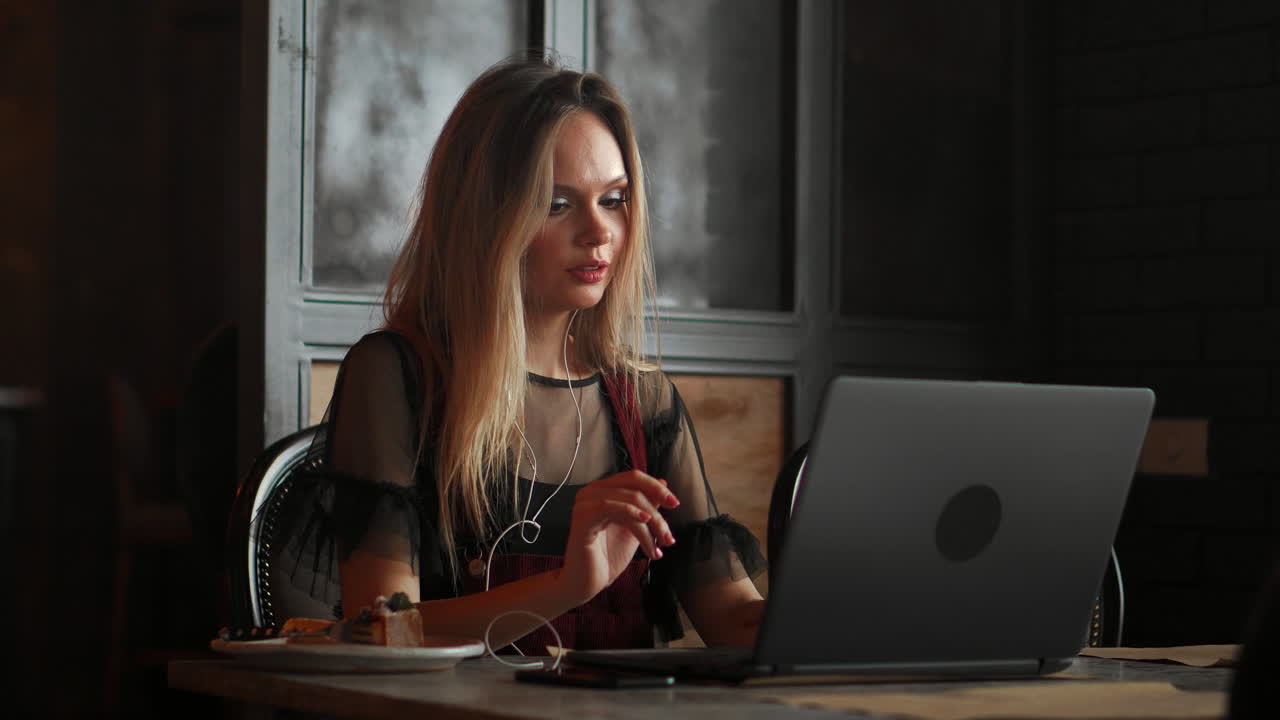 young attractive friendly smiling cheerful woman in a stylish yellow coat deals on the Internet by video call using a headset sitting at a table in a cafe in front of a laptop