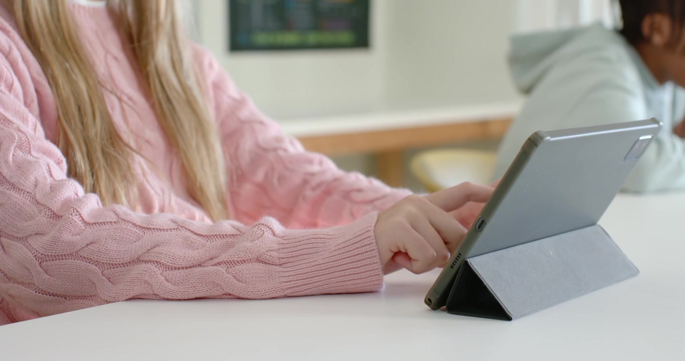 Classmates youth female in pink sweater tapping tablet folio in class while male in hoodie leaning