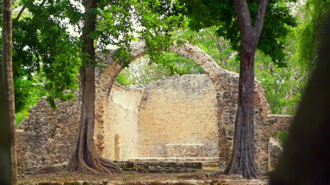 Una toma con inclinación descendente muestra las ruinas históricas de la Capilla Española, una mezcla arquitectónica de estilos maya y colonial, rodeadas de exuberantes árboles.