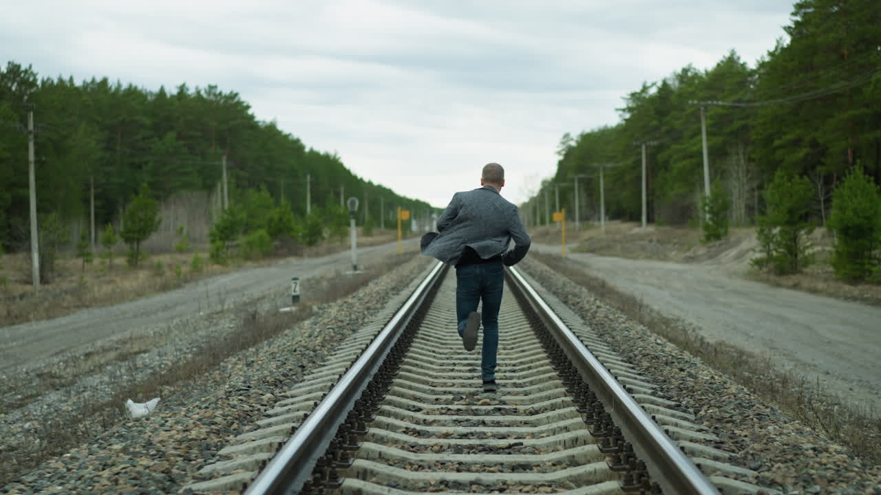 una vista de cerca de un hombre de edad que corre en una vía ferroviaria, él está usando un traje gris, vaqueros, y zapatos de lona, con una vista de árboles y postes eléctricos y otros postes de señales en los alrededores