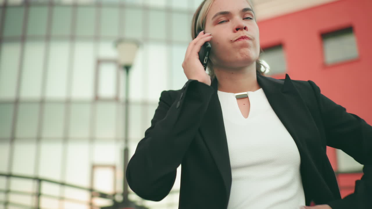 Donna infuriata in abito professionale, in piedi con le braccia conserte mentre è al telefono, con un aspetto visibilmente stanco e turbato in un ambiente urbano moderno con persone sfocate sullo sfondo in lontananza
