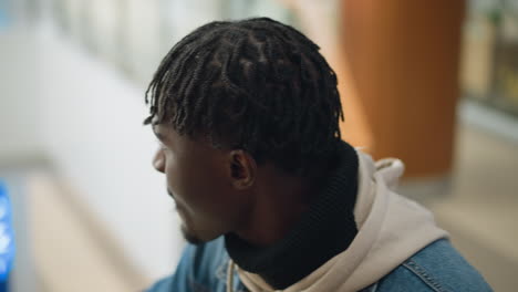 young man in denim jacket and hoodie looking down thoughtfully with focused expression, reflecting in modern indoor space while holding paper cup of coffee